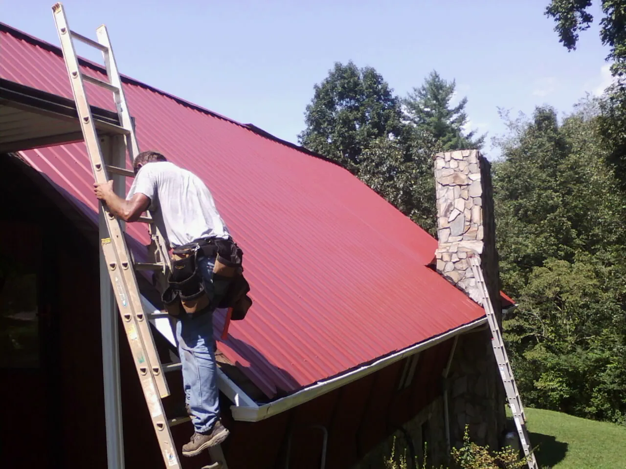 Roofer on a ladder beside a red metal roof and stone chimney — Soffit & Fascia Repair by Roofing Brothers in Santa Ysabel